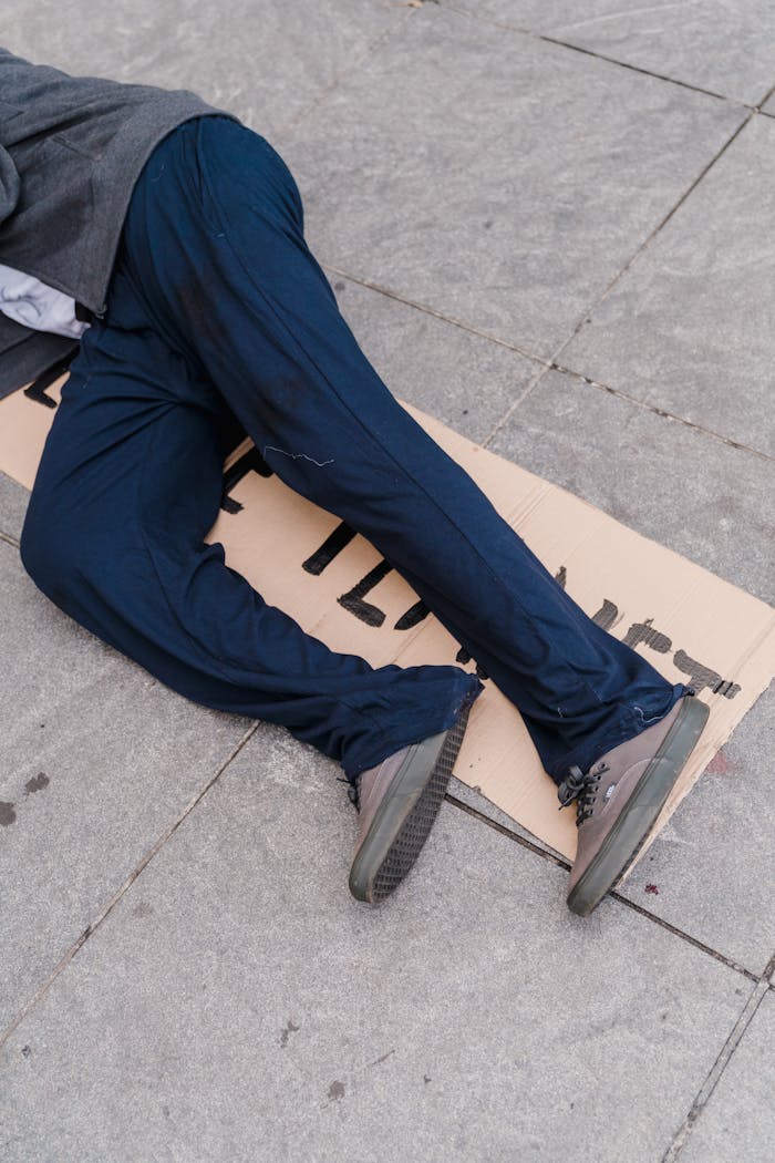 heros-img A person in blue pants lies on a sidewalk with a cardboard sign, symbolizing homelessness.