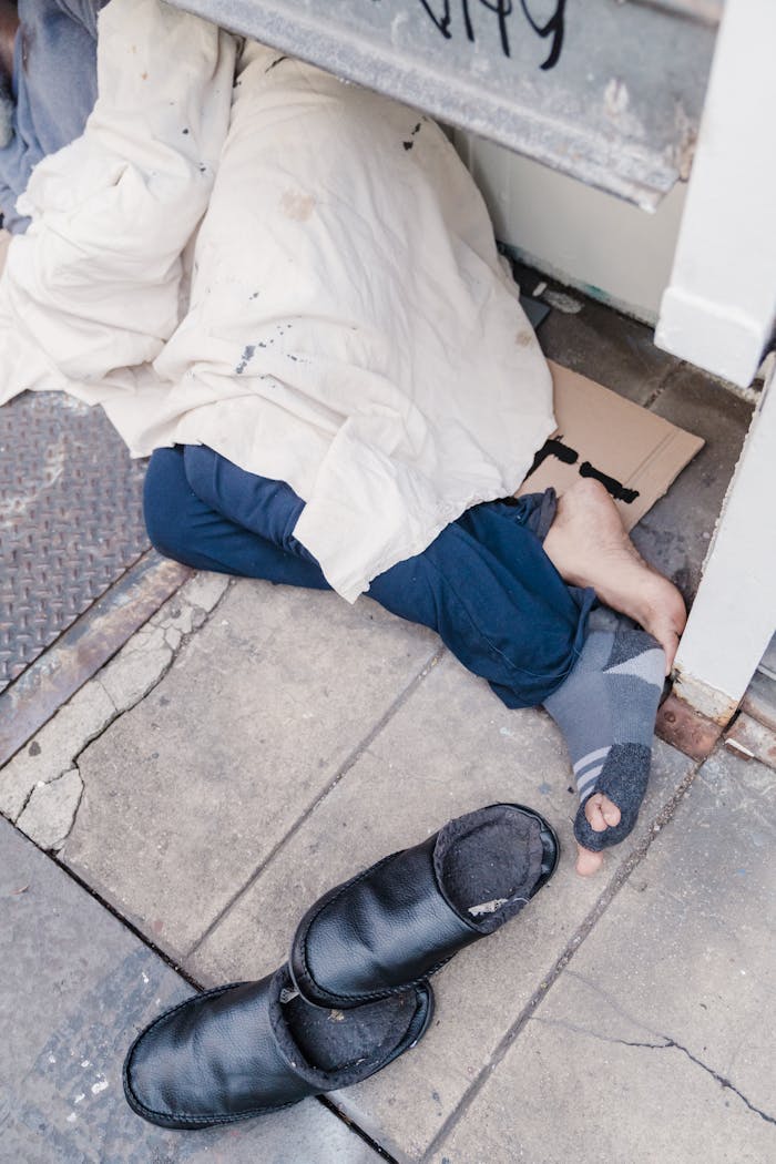 about-us A person sleeps on the sidewalk under a blanket, highlighting homelessness and urban struggles.
